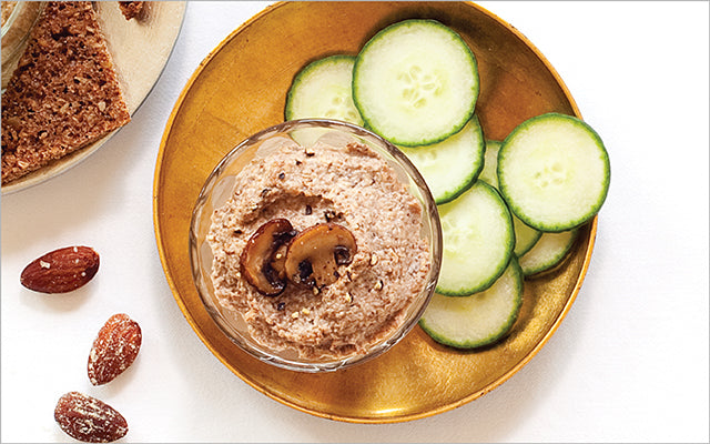 Gold bowl of mushroom pâté topped with a mushroom slice, with cucumber rounds and bread