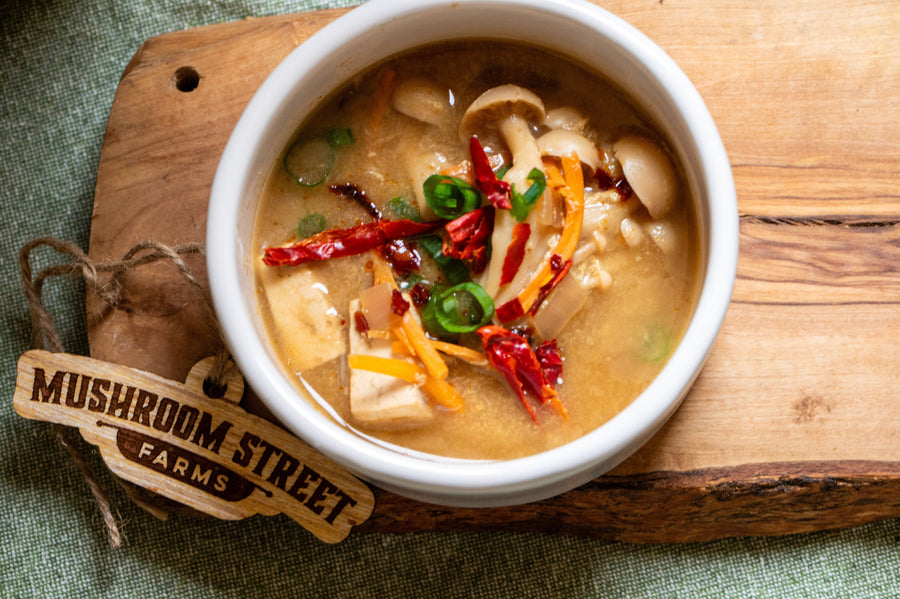White bowl of vegan hot and sour soup with mushrooms and chilli, on a Mushroom Street Farms board