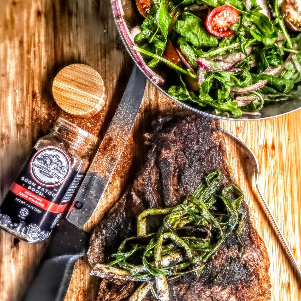 Blackened porterhouse steak on a wooden board with charred green onions, a greens salad and a seasoning jar