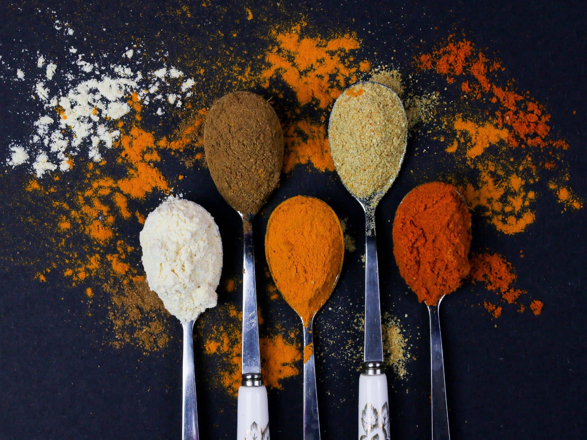 Five spoons holding different coloured spice powders on a dark surface