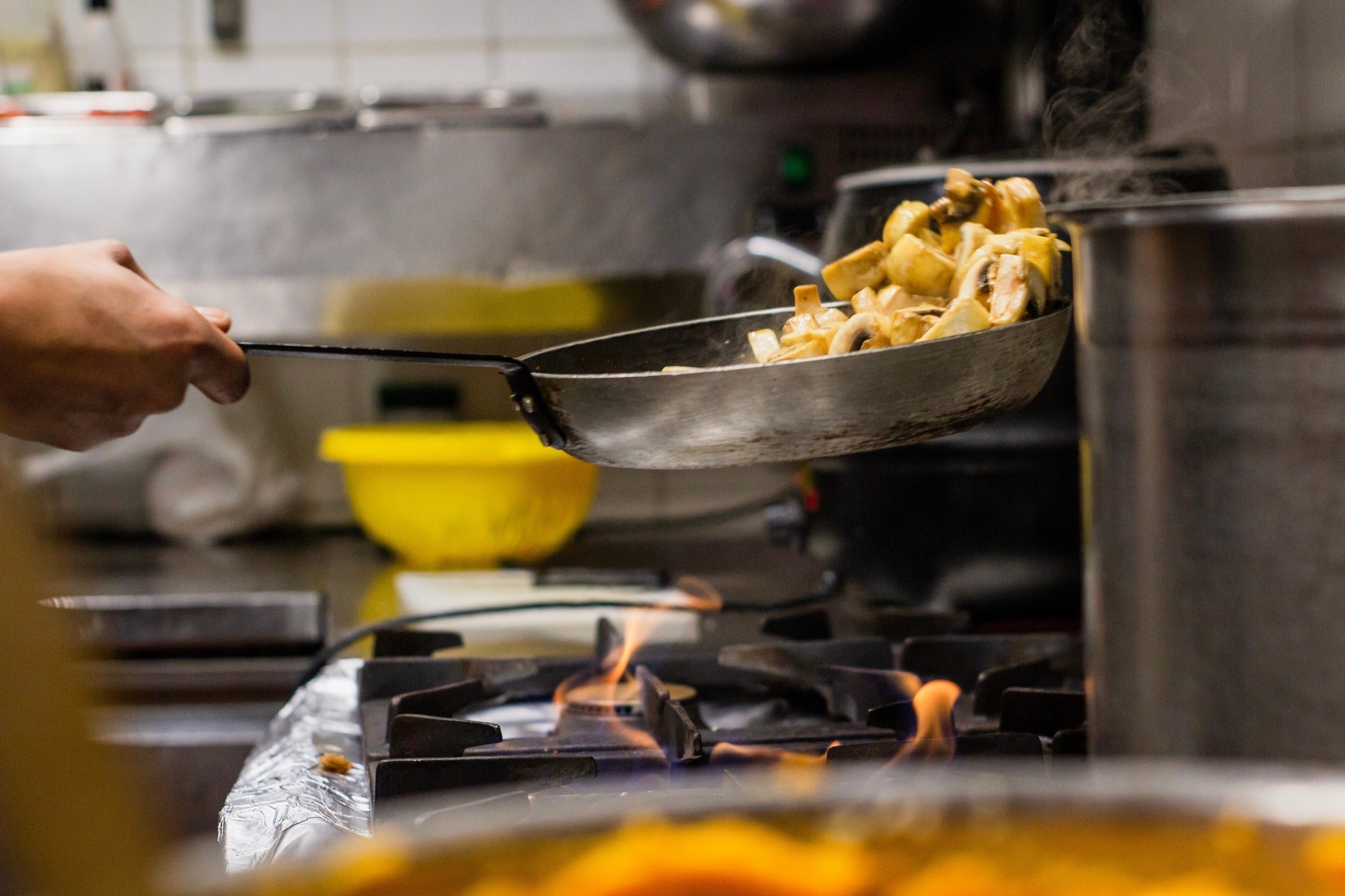 Person in industrial kitchen cooking mushrooms in a pan over fire